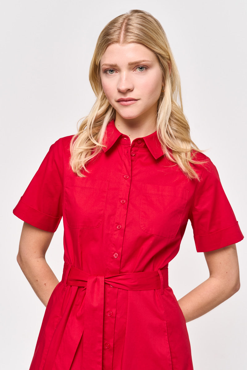 Eine junge Frau mit langen blonden Haaren trägt ein knallrotes, kurzärmeliges Midi-Hemdkleid aus Baumwollpopeline mit Kragen und Bindegürtel von Princess Goes Hollywood. Sie steht vor einem minimalen Hintergrund und schaut leicht ernst, die Arme sind entspannt an ihren Seiten.