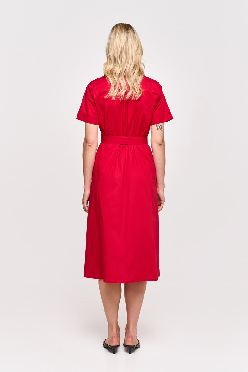 Eine Frau mit gewelltem blondem Haar ist von hinten zu sehen, barfuß, in einem roten Midi-Hemdkleid aus Baumwollpopeline mit einem Taillengürtel von Princess Goes Hollywood. Sie hat eine kleine Tätowierung auf ihrem linken Arm und steht vor einem schlichten weißen Hintergrund.