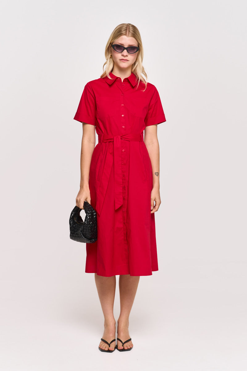 Eine Frau steht vor einem schlichten hellen Hintergrund und trägt ein mittellanges rotes Hemdkleid aus Baumwollpopeline mit passendem Taillengürtel von Princess Goes Hollywood. Sie hat blondes Haar, eine Sonnenbrille, eine schwarze strukturierte Handtasche und schwarze Sandalen.