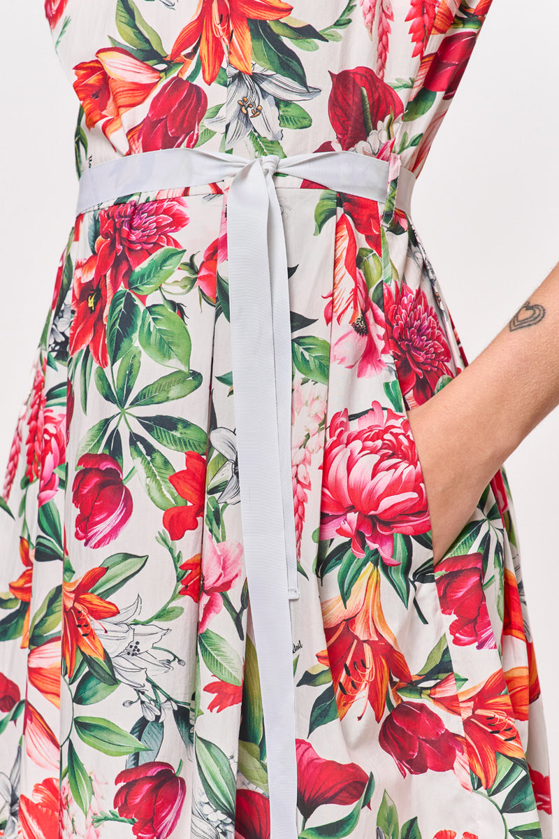Nahaufnahme einer Person, die ein weißes Baumwoll-Minikleid von Princess Goes Hollywood mit einem lebhaften Blumendruck in Rot, Rosa, Grün und Gelb trägt. Das Kleid hat einen Gürtel mit weißem Band, eine Seitentasche mit Handinnenseite und zeigt ein kleines Armtattoo.