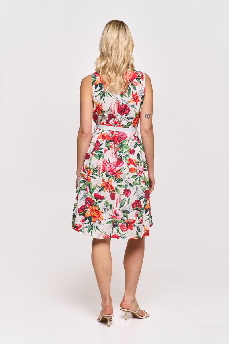 Eine Frau mit gewelltem blondem Haar blickt nach vorne und trägt ein ärmelloses Princess Goes Hollywood Baumwolle Minikleid mit einem leuchtend roten, rosa und grünen Blumendruck. Sie hat ein Schmetterlingstattoo auf ihrem linken Arm und trägt beigefarbene Sandalen mit Absatz auf weißem Grund.