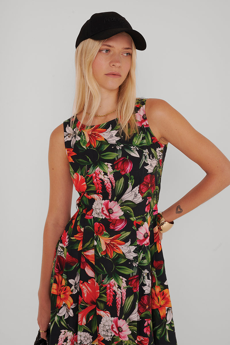 Eine Frau mit glattem blondem Haar trägt eine schwarze Mütze und ein ärmelloses Princess Goes Hollywood Baumwolle Minikleid mit einem leuchtenden tropischen Blumenmuster in Rot, Pink, Orange und Grün. Sie steht vor einem schlichten hellen Hintergrund und zeigt ihr kleines Herz-Tattoo.