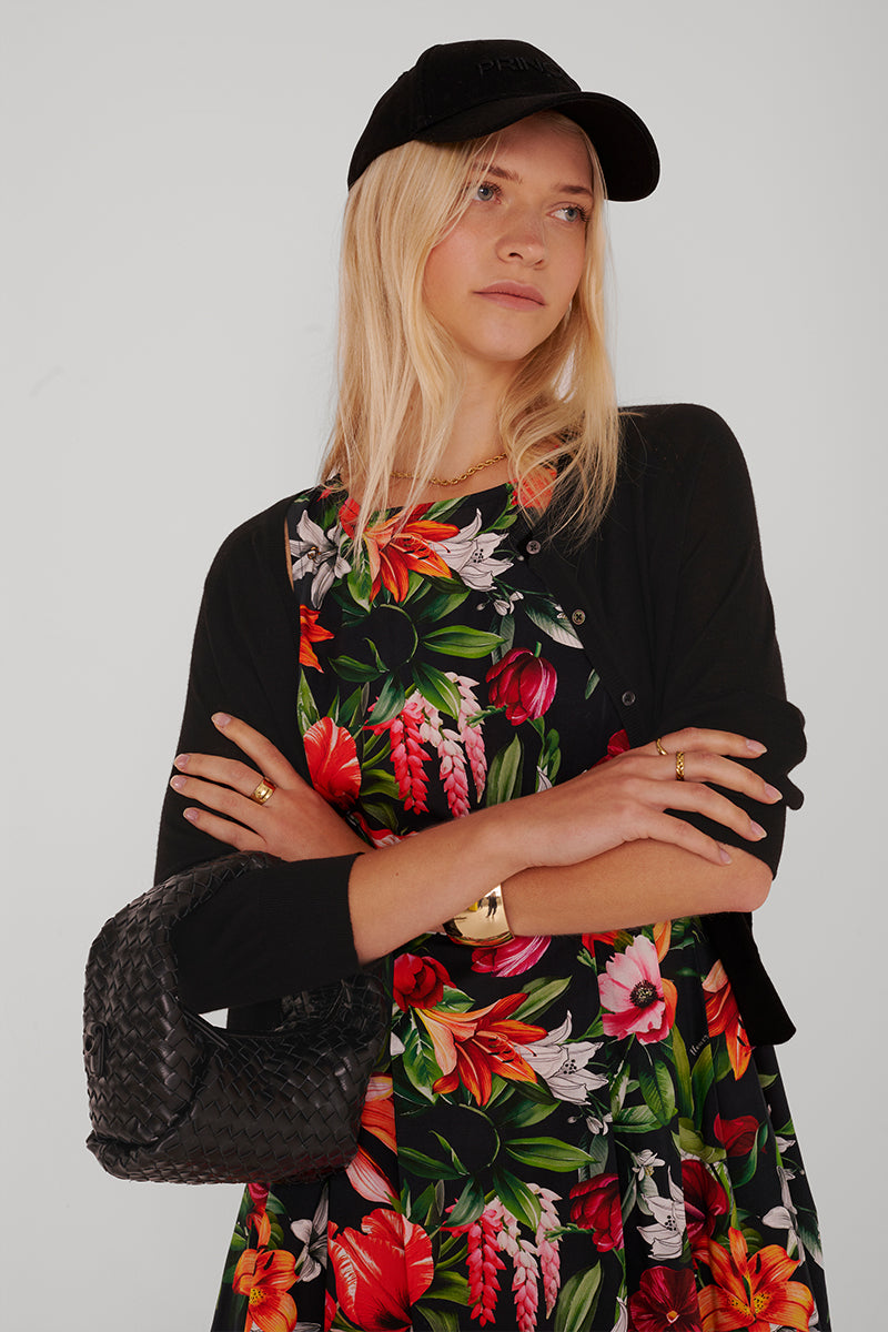 Eine Frau mit langen blonden Haaren trägt eine schwarze Baseballmütze und eine Princess Goes Hollywood-Strickjacke aus Woll-Seide-Gemisch über einem bunten Blumenkleid. Sie trägt eine strukturierte schwarze Handtasche, hat die Arme verschränkt und schaut mit einem neutralen Blick zur Seite.