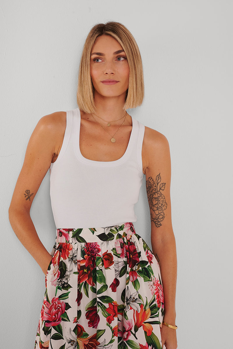 Eine Frau mit blondem Bob trägt ein weißes Basic-Top aus Baumwolle mit Rundhalsausschnitt von Princess Goes Hollywood und einen hoch taillierten geblümten Rock. Florale Tattoos und goldener Schmuck sind sichtbar, sie blickt neutral zur Seite.