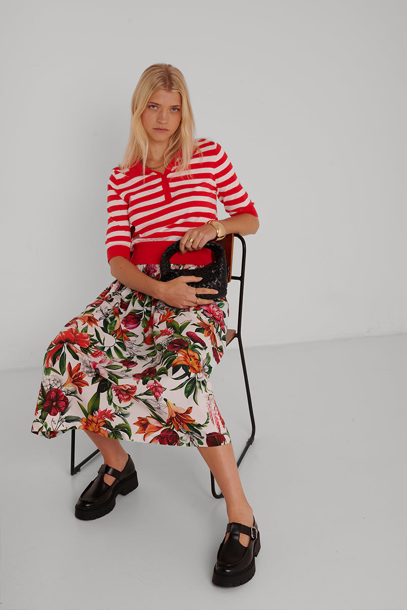 Eine Frau mit blonden Haaren sitzt in einem minimalistischen Raum und trägt ein rot-weiß gestreiftes Top, einen geblümten Midirock aus Baumwolle und schwarze Chunky-Schuhe von Princess Goes Hollywood. Sie hält eine kleine schwarze Handtasche.