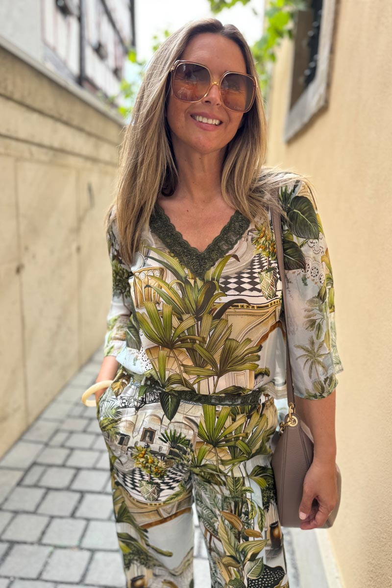 Eine Frau mit langem, glattem blondem Haar lächelt auf einem kopfsteingepflasterten Weg. Sie trägt eine Princess Goes Hollywood Seidenhose mit Safari-Print, elastischem Bund und Spitzenkragen, eine übergroße Sonnenbrille und eine taupefarbene Umhängetasche.