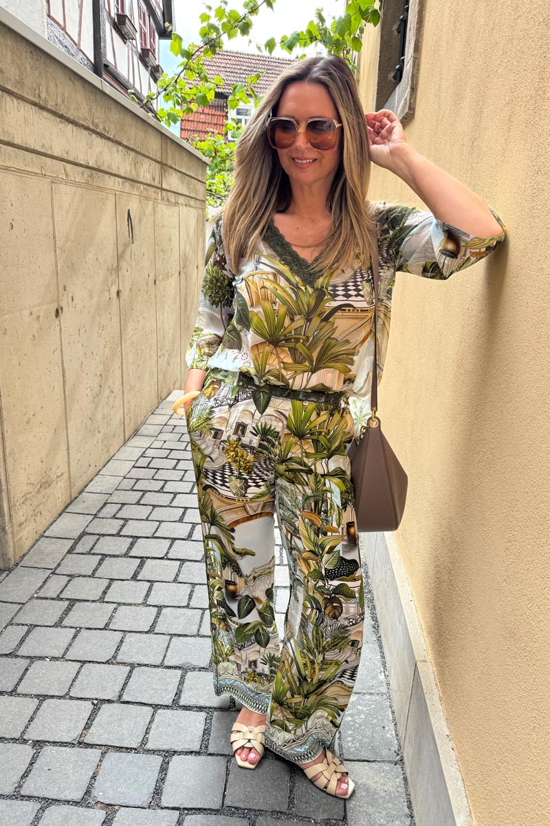 Eine Frau mit langem, hellem Haar lächelt in einer engen Gasse. Sie trägt eine braune Princess Goes Hollywood-Sonnenbrille, ein passendes Oberteil mit Safaridruck und eine Hose mit weitem Bein, elastischem Bund und Taschen. Sie trägt eine taupefarbene Umhängetasche und hat leichte Sandalen an. Beigefarbene Steinmauern umgeben sie.