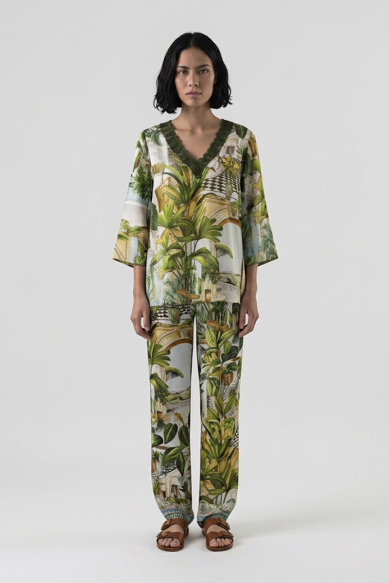 Eine Frau mit schulterlangen dunklen Haaren steht barfuß vor einem unifarbenen Hintergrund und trägt ein lockeres Safari-Print-Set von Princess Goes Hollywood mit tropisch grünen und gelben Palmenmotiven und einer lässigen Seidenhose mit elastischem Bund.