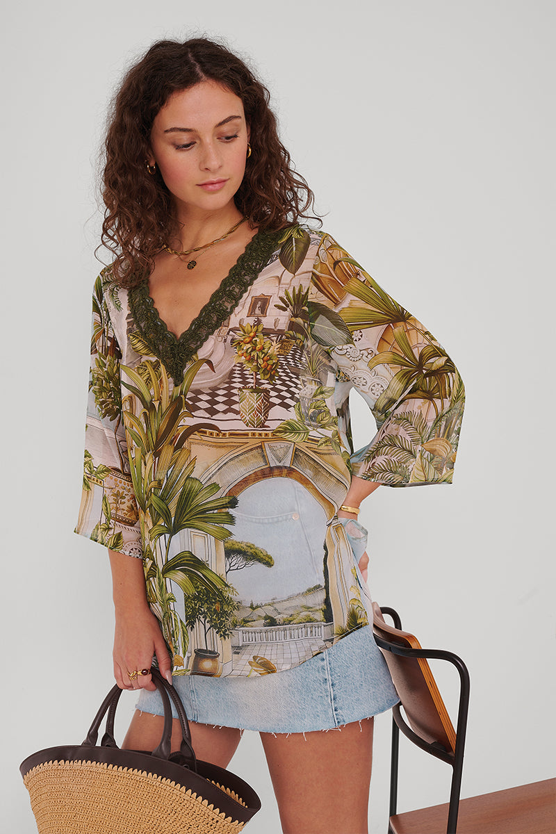 Eine Frau mit lockigen braunen Haaren trägt eine durchsichtige Seidenbluse mit Safaridruck von Princess Goes Hollywood mit einem tiefgrünen V-Ausschnitt und Naturmustern, gepaart mit einem hellblauen Jeansminirock. Sie hält eine geflochtene Strohtasche in der Hand, steht neben einem Holzstuhl und blickt nach links unten.