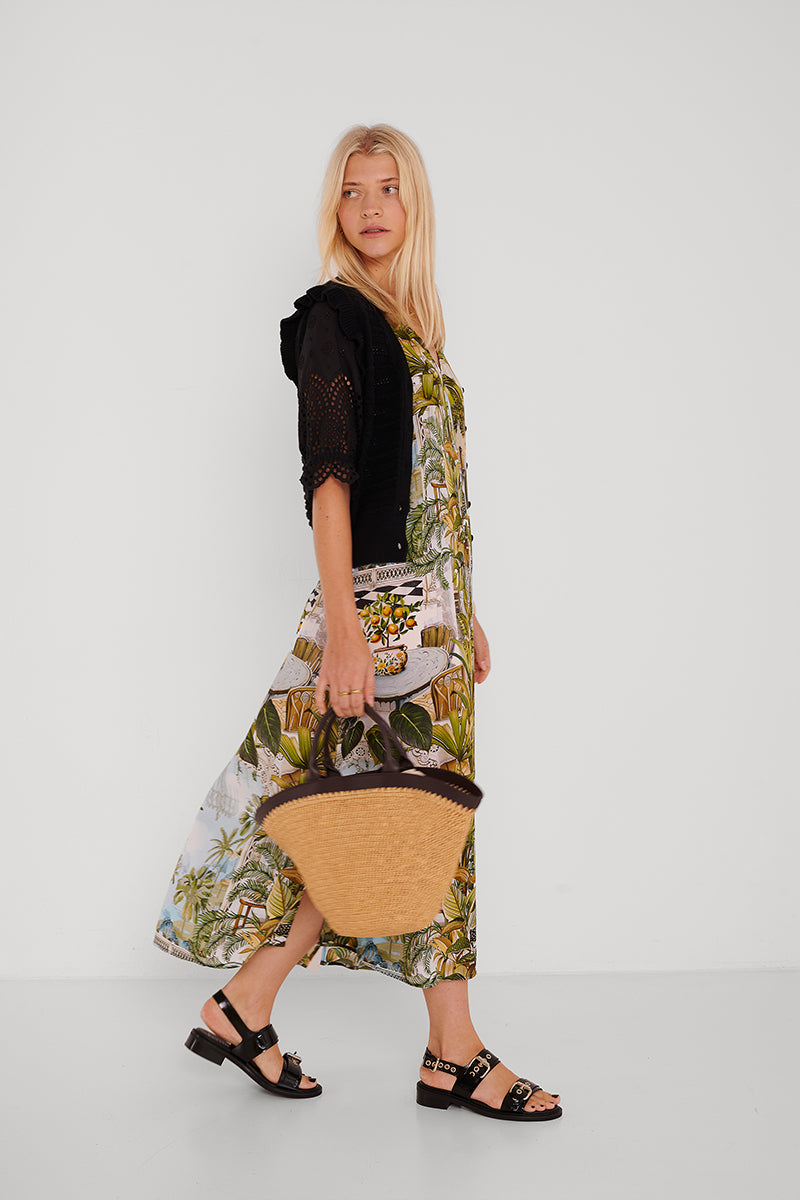 Eine Frau mit langen blonden Haaren trägt eine Princess Goes Hollywood Safari-Print Maxikleid Seide mit V-Ausschnitt, eine schwarze kurzärmelige Häkelstrickjacke, schwarze Sandalen und eine geflochtene Tragetasche aus Stroh vor einem schlichten weißen Hintergrund.