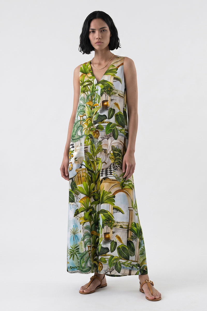 Eine Frau mit kurzen schwarzen Haaren trägt ein ärmelloses Maxikleid mit V-Ausschnitt und Safari-Print von Princess Goes Hollywood, das mit üppigen grünen tropischen und architektonischen Motiven verziert ist. Sie steht vor einem weißen Hintergrund und trägt braune flache Sandalen.
