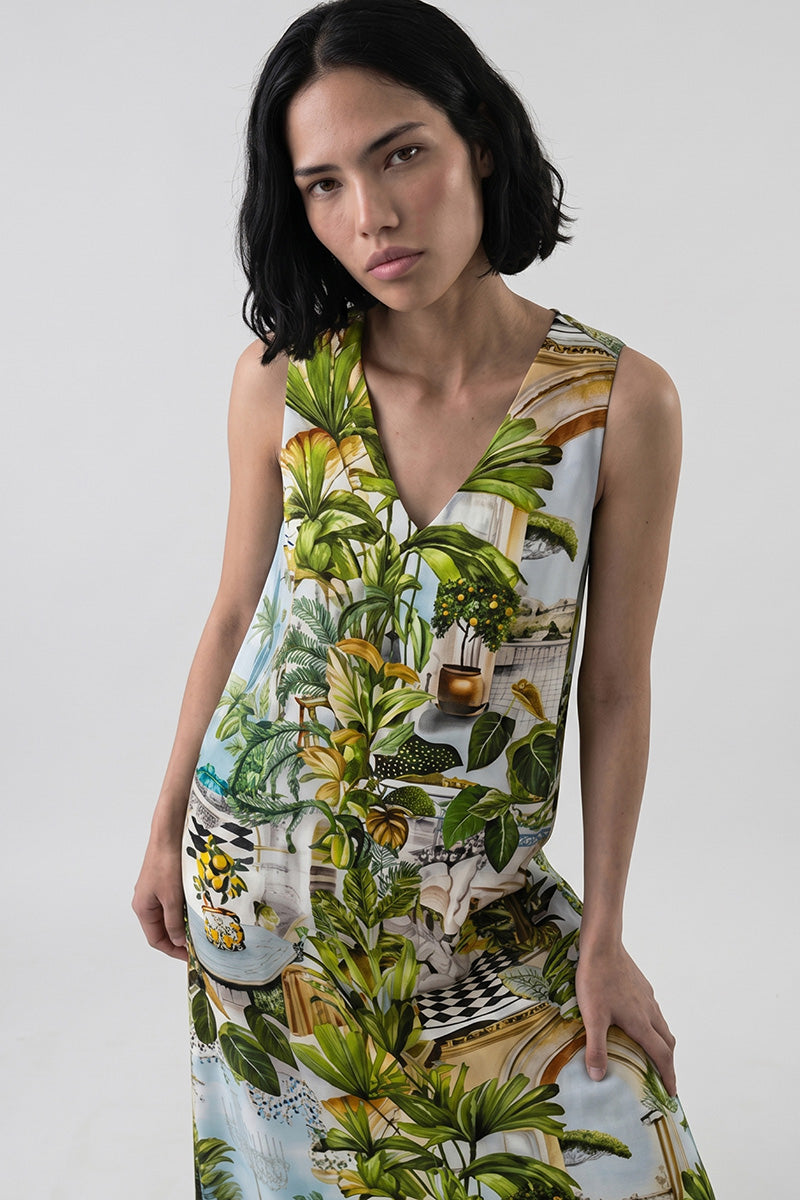 Eine Frau mit mittellangem, dunklem Haar trägt ein Princess Goes Hollywood Seide Maxikleid mit einem lebhaften botanischen und Safaridruck aus grünen Blättern, Pflanzen und dekorativen Motiven. Sie steht vor einem hellgrauen Hintergrund und blickt mit einem neutralen Blick nach oben.