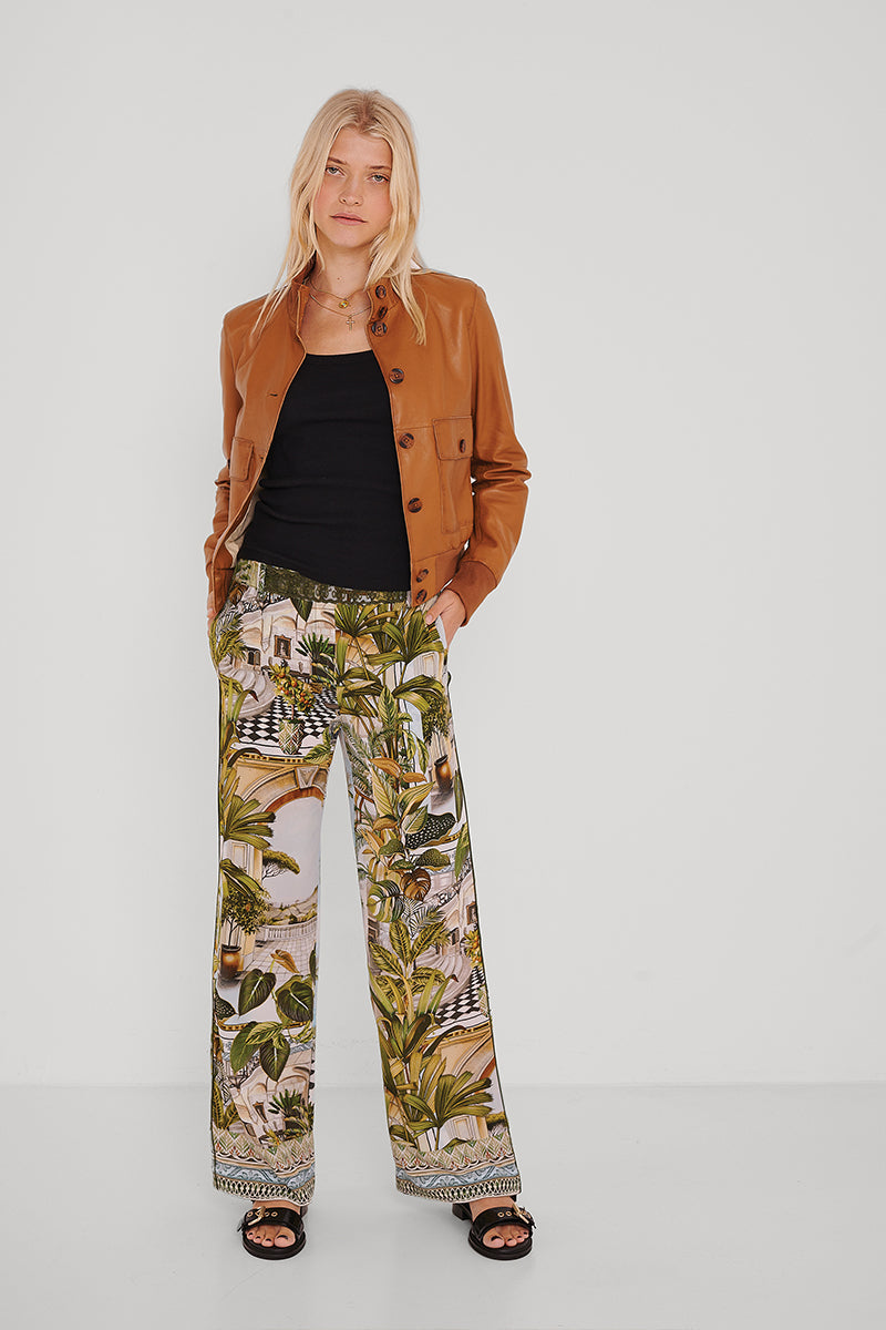 Eine Frau mit langen blonden Haaren steht vor einem weißen Hintergrund und trägt eine braune Lederjacke von Princess Goes Hollywood über einem schwarzen Baumwolloberteil mit Rundhalsausschnitt, eine weite Hose mit grünem und braunem Tropenmuster und schwarze Sandalen. Ihre Hände stecken in den Taschen.