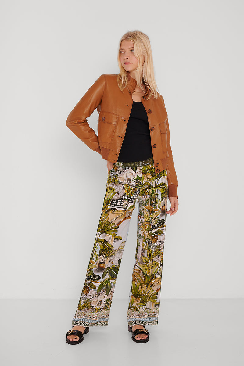 Eine Frau mit glattem blondem Haar trägt eine braune Lederjacke über einem schwarzen Oberteil und eine hoch taillierte Princess Goes Hollywood Seidenhose mit grünem tropischem Safari-Print, gepaart mit schwarzen Slide-Sandalen, und steht vor einer schlichten weißen Wand.