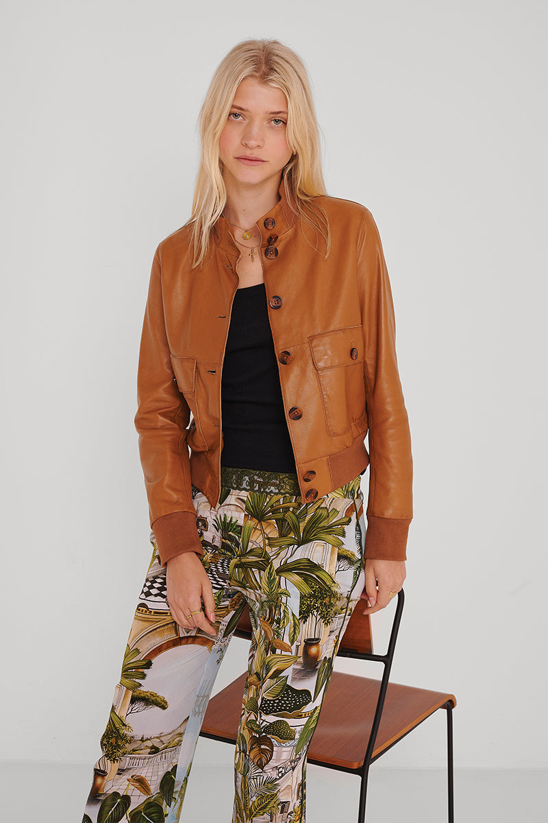 Eine Frau mit langen blonden Haaren trägt eine hellbraune Lederjacke über einem schwarzen Oberteil und eine Seidenhose mit elastischem Bund im Safaridruck von Princess Goes Hollywood. Sie steht vor einem einfachen Stuhl vor einem schlichten weißen Hintergrund und schaut ruhig in die Kamera.