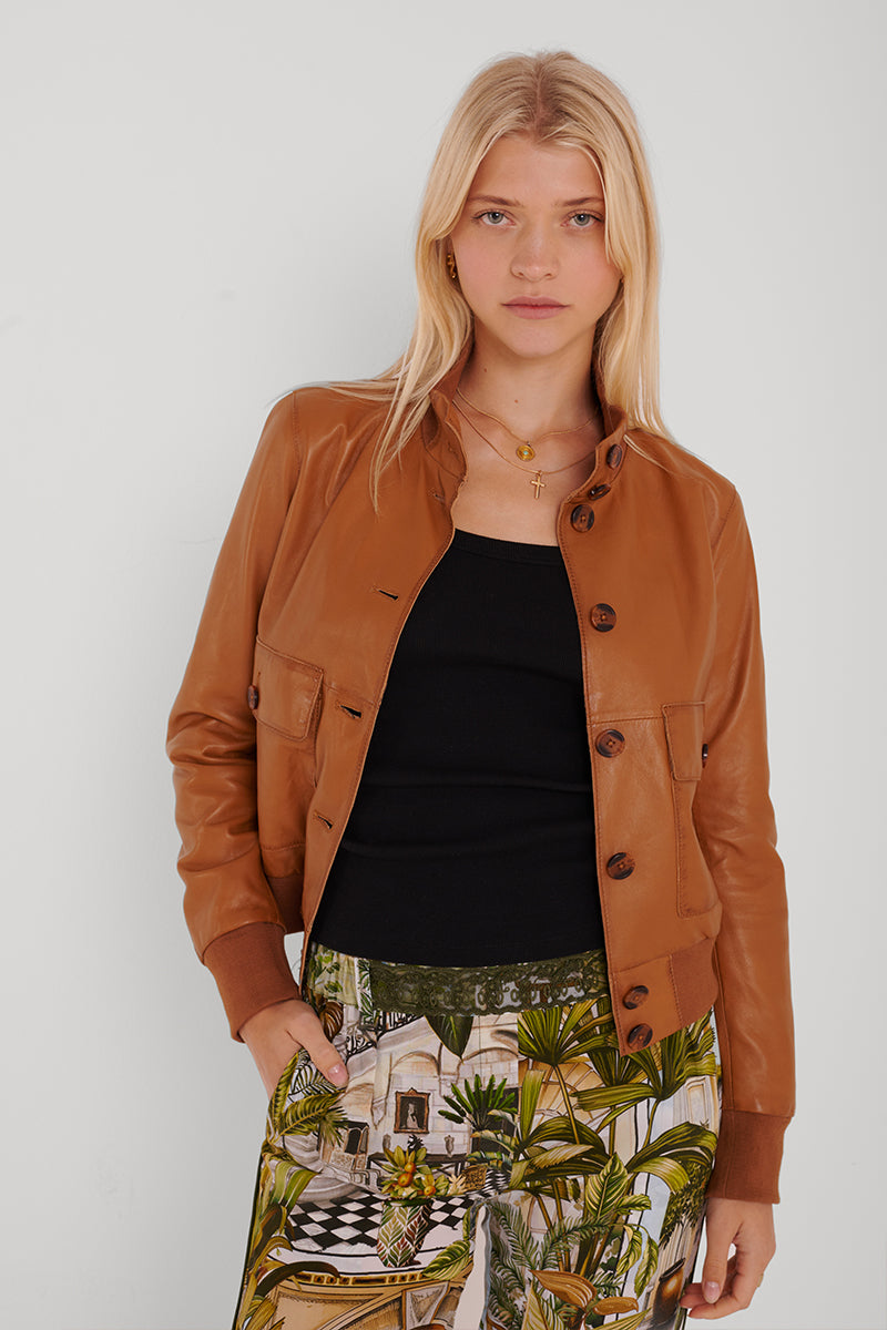 Eine Frau mit langen blonden Haaren trägt eine hellbraune Lederjacke mit Knopfleiste von Princess Goes Hollywood über einem schwarzen Rundhals-Top und einer Hose mit grünem und braunem Tropenprint. Sie steht vor einem weißen Hintergrund, die Hände in den Taschen, und schaut in die Kamera.