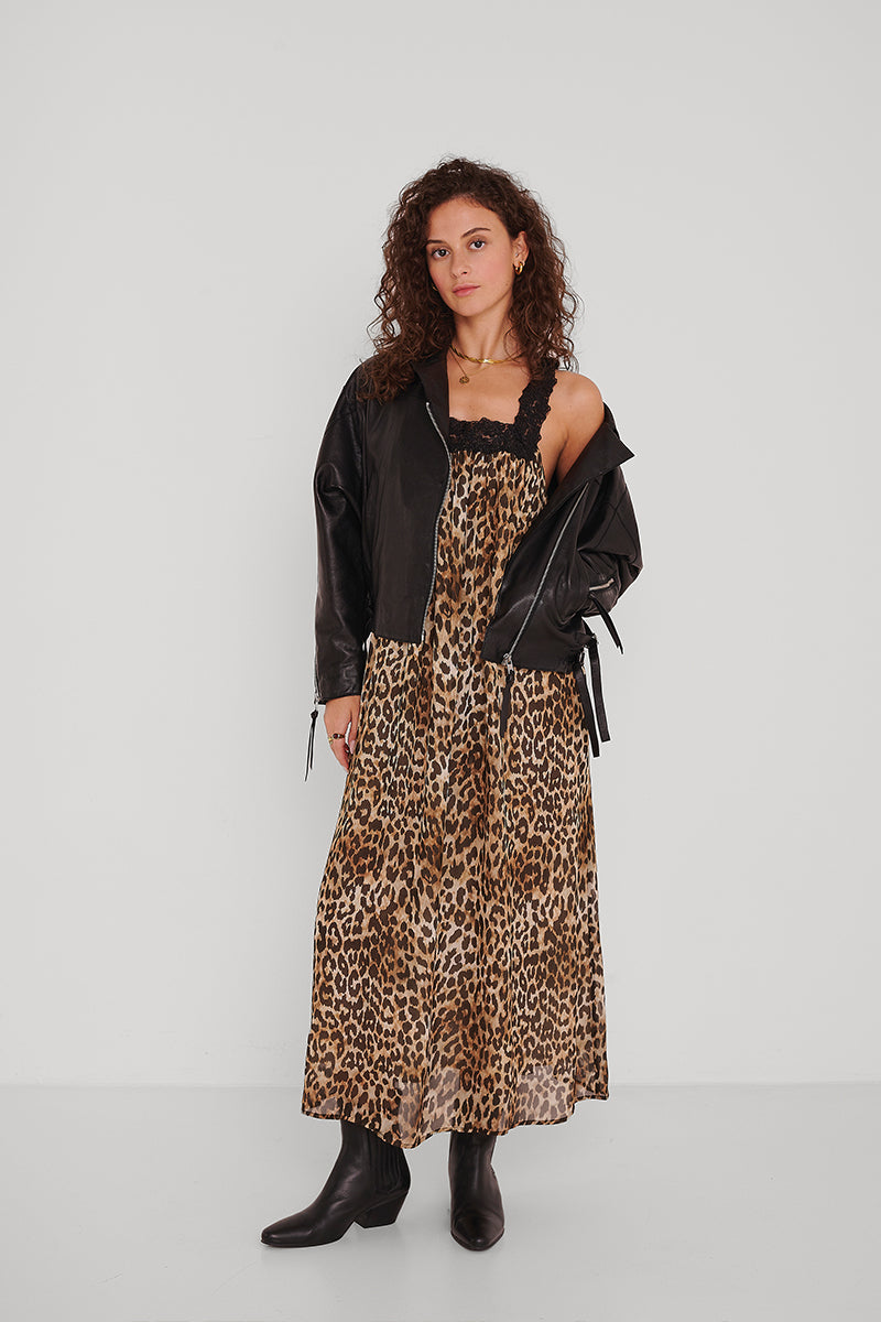 Eine Frau mit lockigem Haar trägt eine schwarze Lederjacke von Princess Goes Hollywood, ein Maxikleid mit Leopardenmuster und Spitzenbesatz sowie schwarze Stiefel. Sie steht vor einer schlichten weißen Wand und schaut selbstbewusst in die Kamera.
