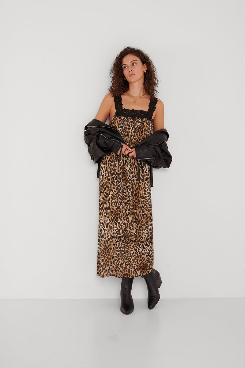 Eine Frau mit lockigem Haar lehnt an einer weißen Wand. Sie trägt ein Maxikleid im Leopardenmuster von Princess Goes Hollywood mit schwarzem Spitzenbesatz, eine schwarze Lederjacke über den Schultern und schwarze Ankle Boots. Sie blickt in einer entspannten Pose sanft zur Seite.