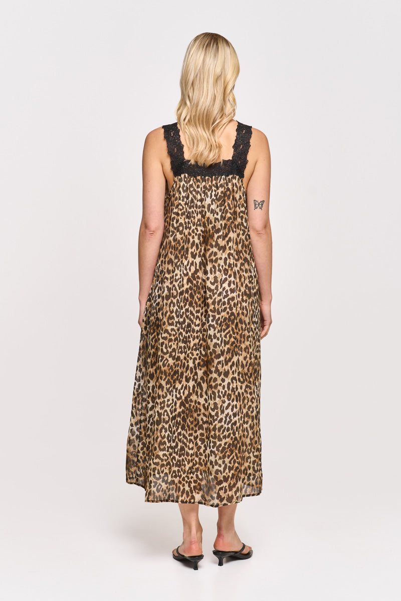 Eine Frau mit hellblonden Wellen steht mit dem Gesicht nach hinten auf einem weißen Hintergrund. Sie trägt ein ärmelloses, wadenlanges Princess Goes Hollywood Leoprint Maxikleid mit schwarzem Spitzen-Träger, schwarze Sandalen und hat ein kleines Schmetterlingstattoo auf dem linken Oberarm.