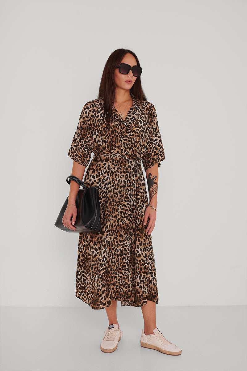 Eine Frau mit langen braunen Haaren und einer sichtbaren Unterarmtätowierung steht vor einem weißen Hintergrund und trägt ein mittellanges, kurzärmeliges Princess Goes Hollywood Leoprint Hemdblusenkleid, weiße Turnschuhe, eine dunkle Sonnenbrille und eine schwarze Handtasche.
