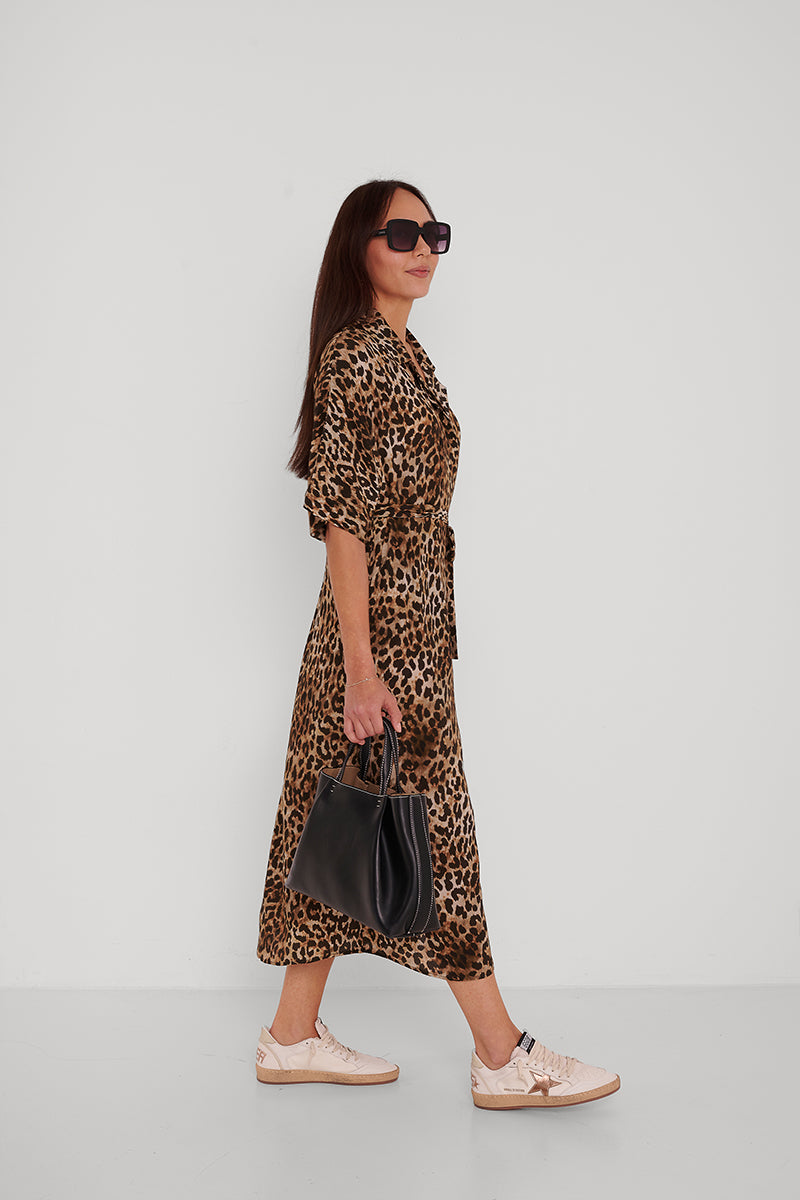 Eine Frau mit langen braunen Haaren, Sonnenbrille und einem Princess Goes Hollywood Leoprint Hemdblusenkleid mit Taillengürtel geht seitwärts vor einem weißen Hintergrund, trägt eine schwarze Handtasche und helle Turnschuhe.