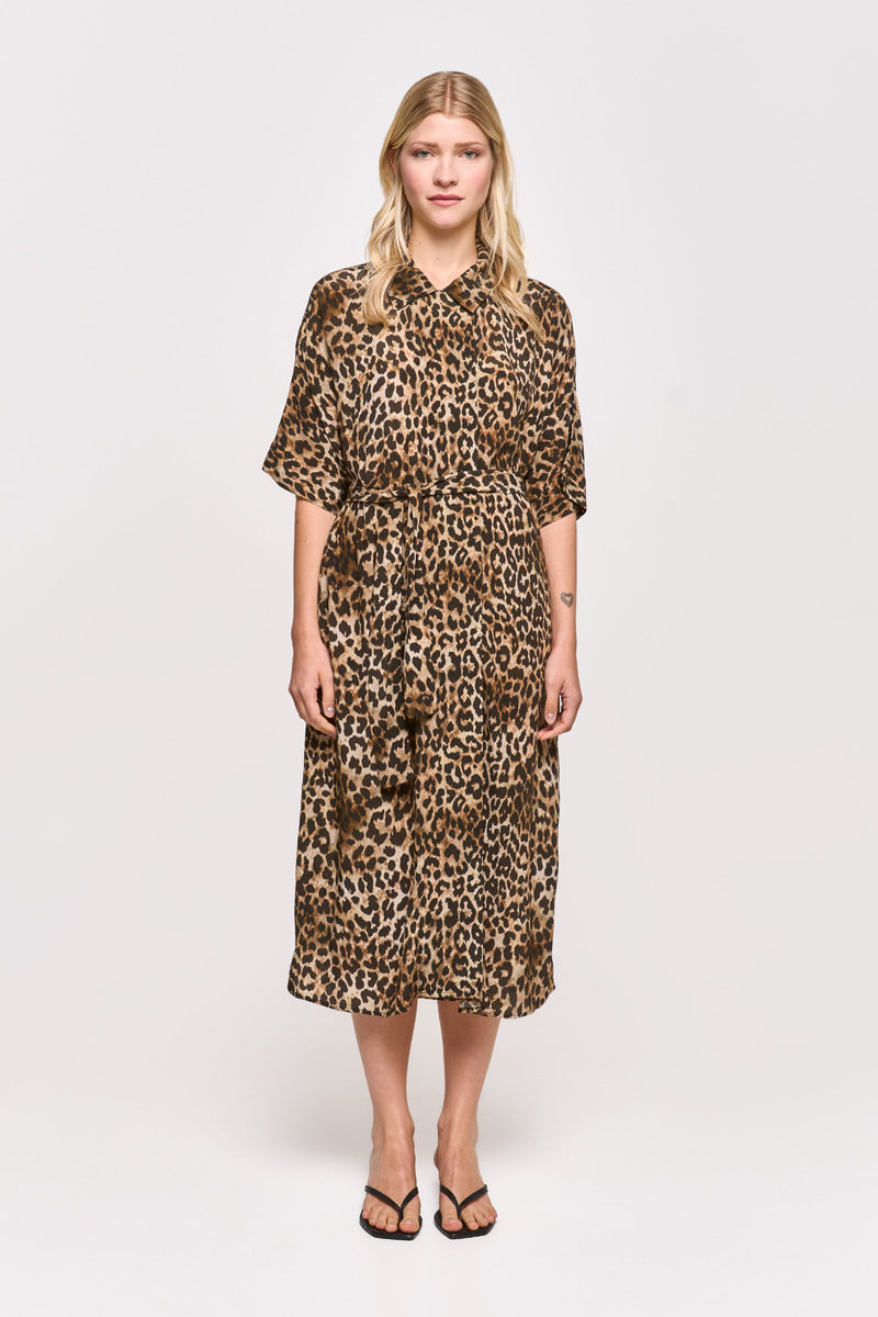 Eine Frau blickt vor einem schlichten weißen Hintergrund nach vorne und trägt ein knielanges Kleid mit Leopardenmuster und einem Taillengürtel von Princess Goes Hollywood. Sie hat glattes, lockeres blondes Haar, schwarze Flip-Flops und ihre Arme ruhen ganz natürlich an ihren Seiten.