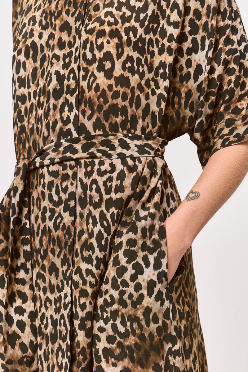 Eine Nahaufnahme einer Person, die ein braun-schwarzes Leoprint Hemdblusenkleid mit Taillengürtel von Princess Goes Hollywood trägt. Eine Hand liegt in der Seitentasche, ein kleines Tattoo am Unterarm; der Hintergrund ist schlicht und hell.
