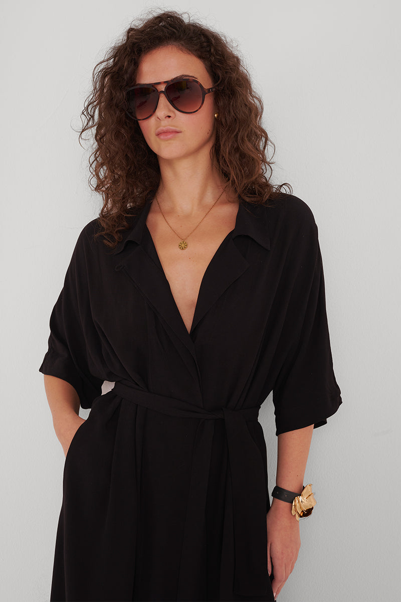 Eine Frau mit lockigem braunem Haar trägt eine große Sonnenbrille, ein Princess Goes Hollywood schwarzes Hemdblusenkleid mit Taillengürtel und tiefem V-Ausschnitt, dazu Goldschmuck und eine schwarze Uhr. Sie steht vor einem hellen Hintergrund und blickt selbstbewusst in die Kamera.