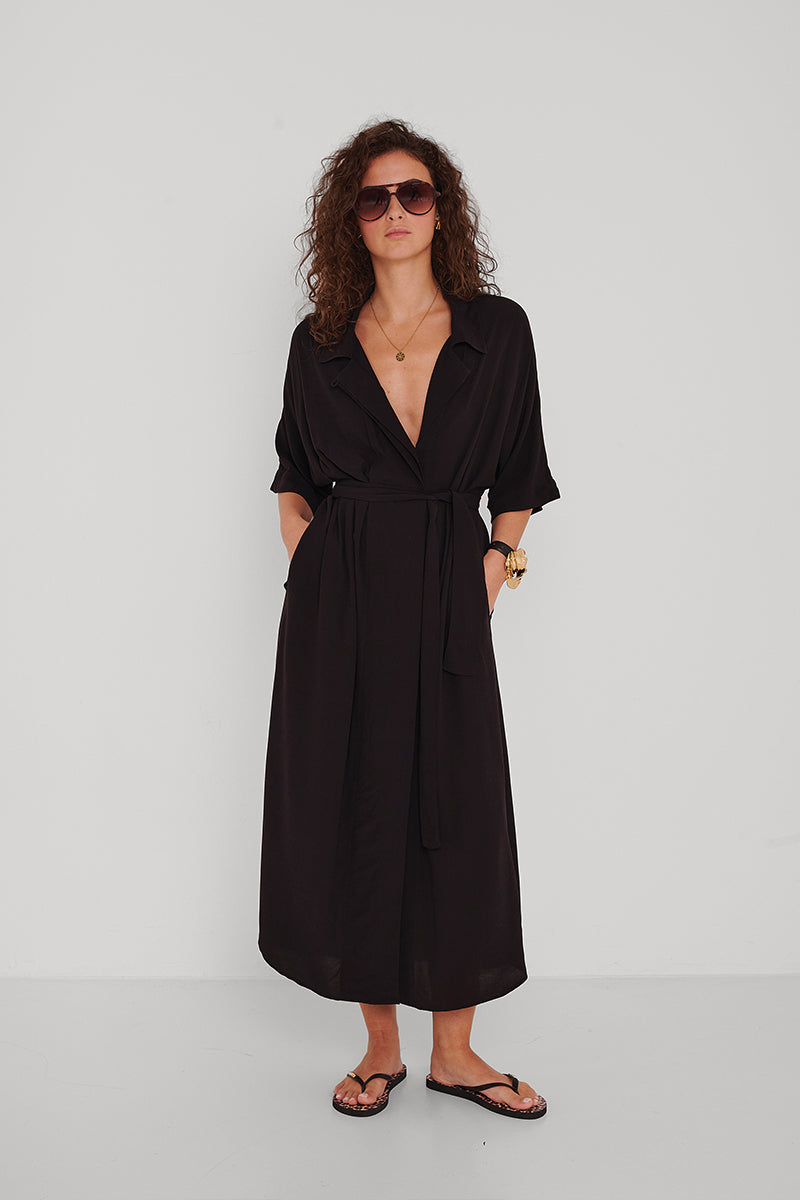 Eine Frau mit lockigem Haar trägt eine dunkle Sonnenbrille, ein schwarzes Princess Goes Hollywood Hemdblusenkleid mit Taillengürtel und Seitentaschen, schwarze Flip-Flops, eine goldene Armbanduhr und eine runde goldene Halskette und steht mit den Händen in den Taschen vor einer weißen Wand.