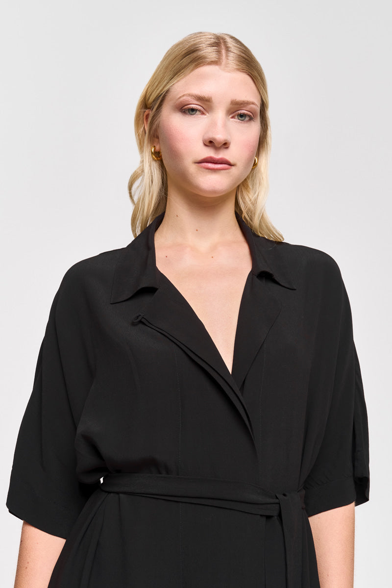 Eine hellhäutige Frau mit langen blonden Haaren trägt ein schwarzes Princess Goes Hollywood-Hemdkleid mit Taillengürtel und Seitentaschen, goldene Ohrringe und posiert vor einem schlichten hellgrauen Hintergrund mit einem neutralen Gesichtsausdruck.