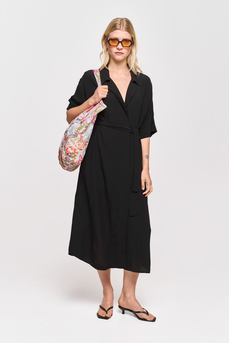Eine Frau mit blondem Bubikopf steht vor einem schlichten weißen Hintergrund. Sie trägt ein schwarzes Kleid mit Gürtel und schwarze Flip-Flops von Princess Goes Hollywood, eine orangefarbene Sonnenbrille und eine bunte Einkaufstasche, die sie über der Schulter recycelt.