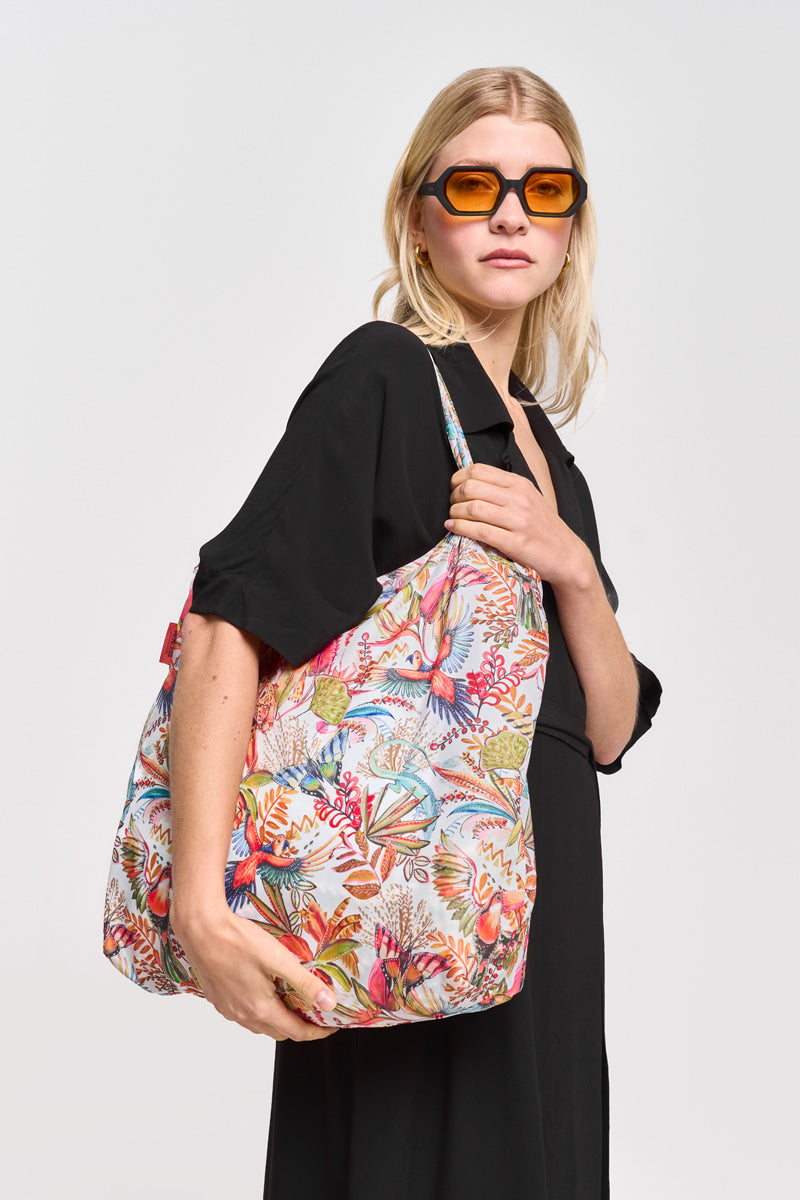 Eine blonde Frau mit bernsteinfarbener Sonnenbrille und schwarzem Princess Goes Hollywood Hemdblusenkleid steht seitlich und schaut in die Kamera. Sie trägt eine farbenfrohe Tasche mit lebhaften tropischen Vogel- und Blumendrucken über der Schulter vor einem schlichten weißen Hintergrund.