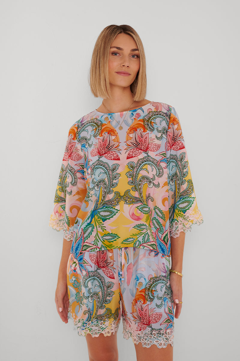 Eine Frau mit einem stumpfen, schulterlangen blonden Haarschnitt steht vor einem weißen Hintergrund und lächelt leicht in einer lockeren Seidenbluse von Princess Goes Hollywood und passenden Shorts mit Paisley-Print, Blumen- und Blattmotiven, Spitzenbordüren und zarten Stickereien an Ärmeln und Saum.