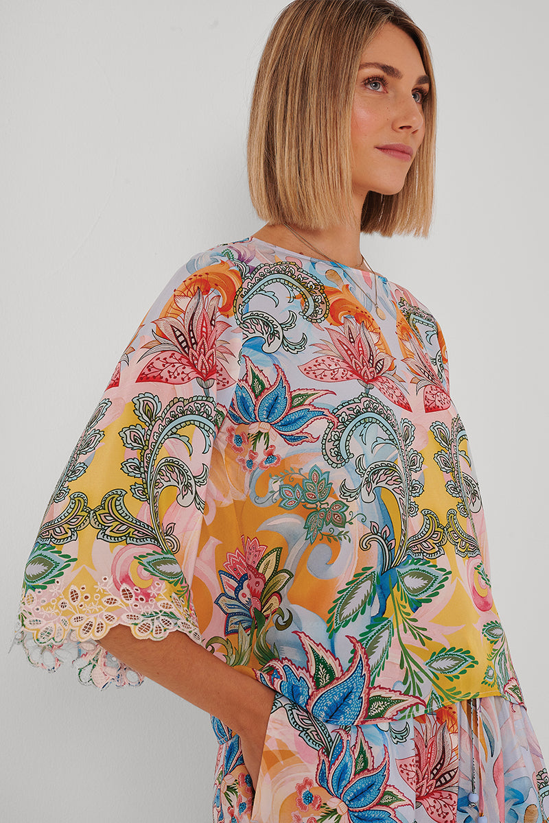 Eine Frau mit glattem blondem Bob trägt eine durchsichtige Princess Goes Hollywood Seidenbluse mit weiten Ärmeln, die mit einem leuchtenden rosa, gelben, blauen und grünen Paisley-Blumenprint und Stickereirändern verziert ist. Sie steht vor einer weißen Wand.