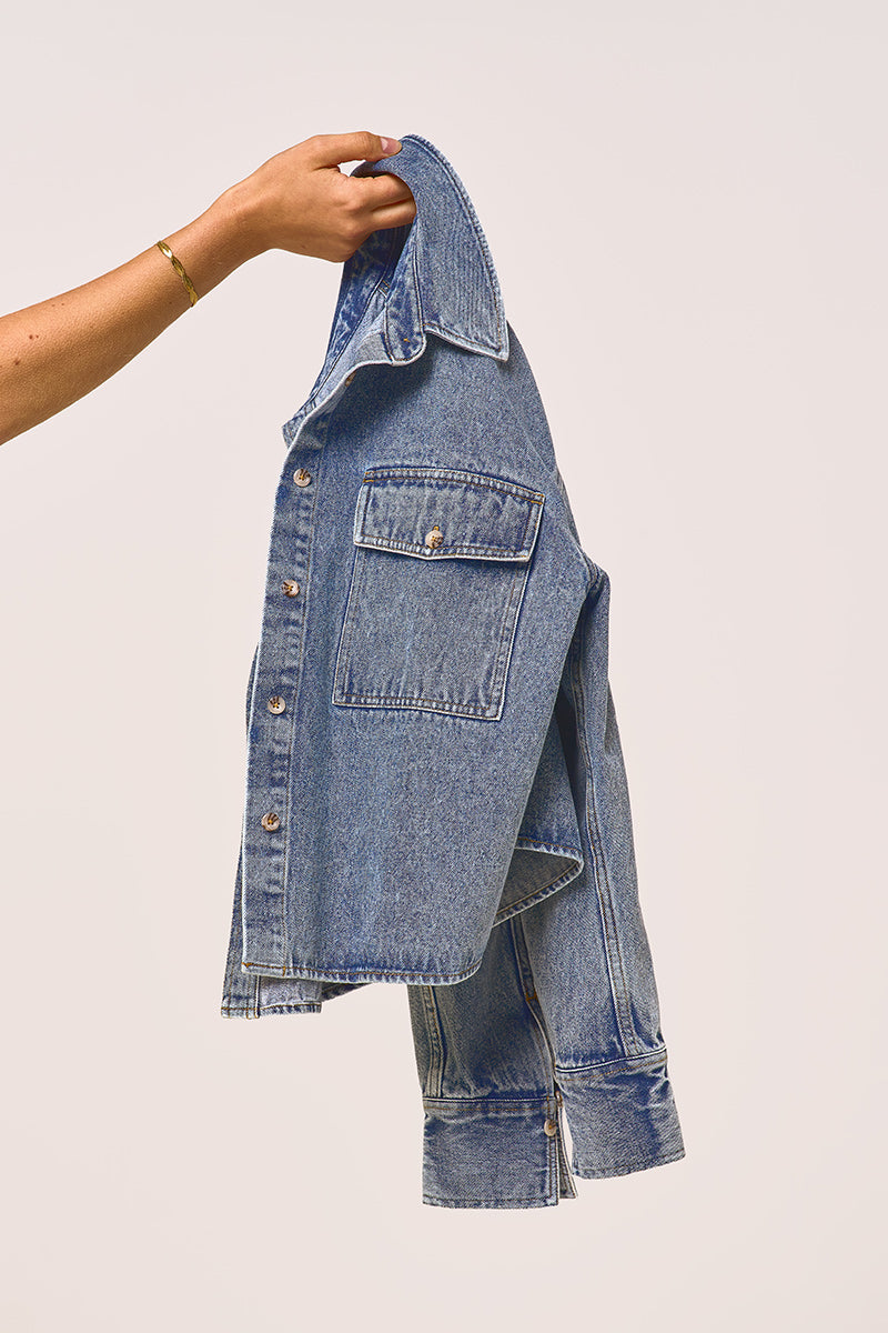 Eine Hand, die ein dünnes Goldarmband trägt, hält eine Princess Goes Hollywood Damen Jeansjacke am Kragen vor einem schlichten hellen Hintergrund hoch. Die blaue Jacke hat Metallknöpfe und eine Brusttasche mit Klappe, Ärmel und Saum hängen natürlich herunter.