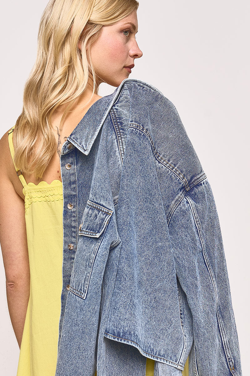 Eine Frau mit blondem, gewelltem Haar trägt ein gelbes Spaghettiträgerkleid und hat sich eine übergroße Princess Goes Hollywood-Jeansjacke über die Schulter gelegt. Sie blickt zur Seite und schaut nach links vor einem schlichten, hellen Hintergrund.
