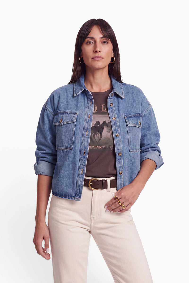 Eine Frau mit glattem, dunklem Haar trägt eine übergroße Damen-Jeansjacke über einem T-Shirt mit Pferdemotiv, eine cremefarbene Hose mit hoher Taille, einen braunen Gürtel und goldene Creolen. Sie steht selbstbewusst mit einer Hand an der Seite vor einem schlichten weißen Hintergrund.