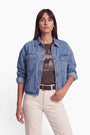 Eine Frau mit glattem, dunklem Haar trägt eine übergroße Damen-Jeansjacke über einem T-Shirt mit Pferdemotiv, eine cremefarbene Hose mit hoher Taille, einen braunen Gürtel und goldene Creolen. Sie steht selbstbewusst mit einer Hand an der Seite vor einem schlichten weißen Hintergrund.