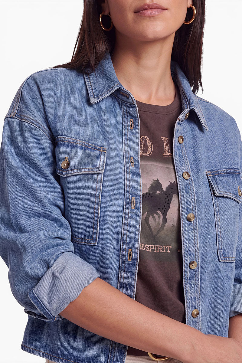 Eine Frau mit braunen Haaren trägt eine übergroße Jeansjacke aus hellblauem Denim über einem schwarzen T-Shirt mit Aufdruck. Die Ärmel der Jacke sind hochgekrempelt, und sie trägt goldene Creolen. Auf dem T-Shirt sind ein Pferd und ein Schriftzug im Western-Stil zu sehen. Ihr Gesicht ist teilweise zu sehen.