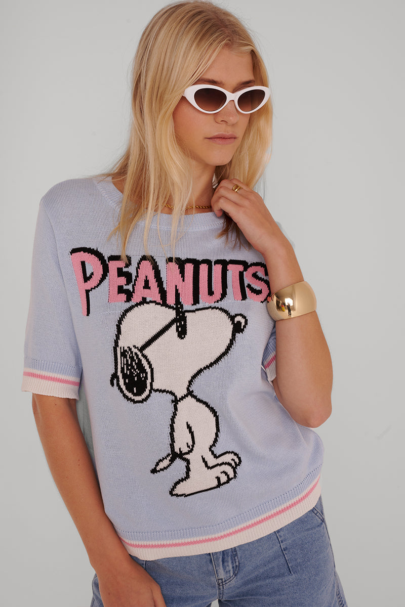 Eine Frau mit langen blonden Haaren trägt blaue Jeans, ein klobiges goldenes Armband und eine weiße Katzenaugen-Sonnenbrille. Sie trägt einen hellblauen Princess Goes Hollywood Peanuts-Pullover mit rosa PEANUTS-Schriftzug und einem großen Snoopy-Motiv auf grauem Hintergrund.