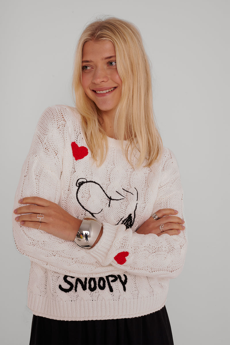 Eine lächelnde Frau mit langen blonden Haaren trägt einen weißen Zopfstrickpullover aus Baumwollmischung mit Snoopy-Stickerei, roten Herzen und "Snoopy"; sie verschränkt die Arme und trägt silberne Ringe vor einem grauen Hintergrund.