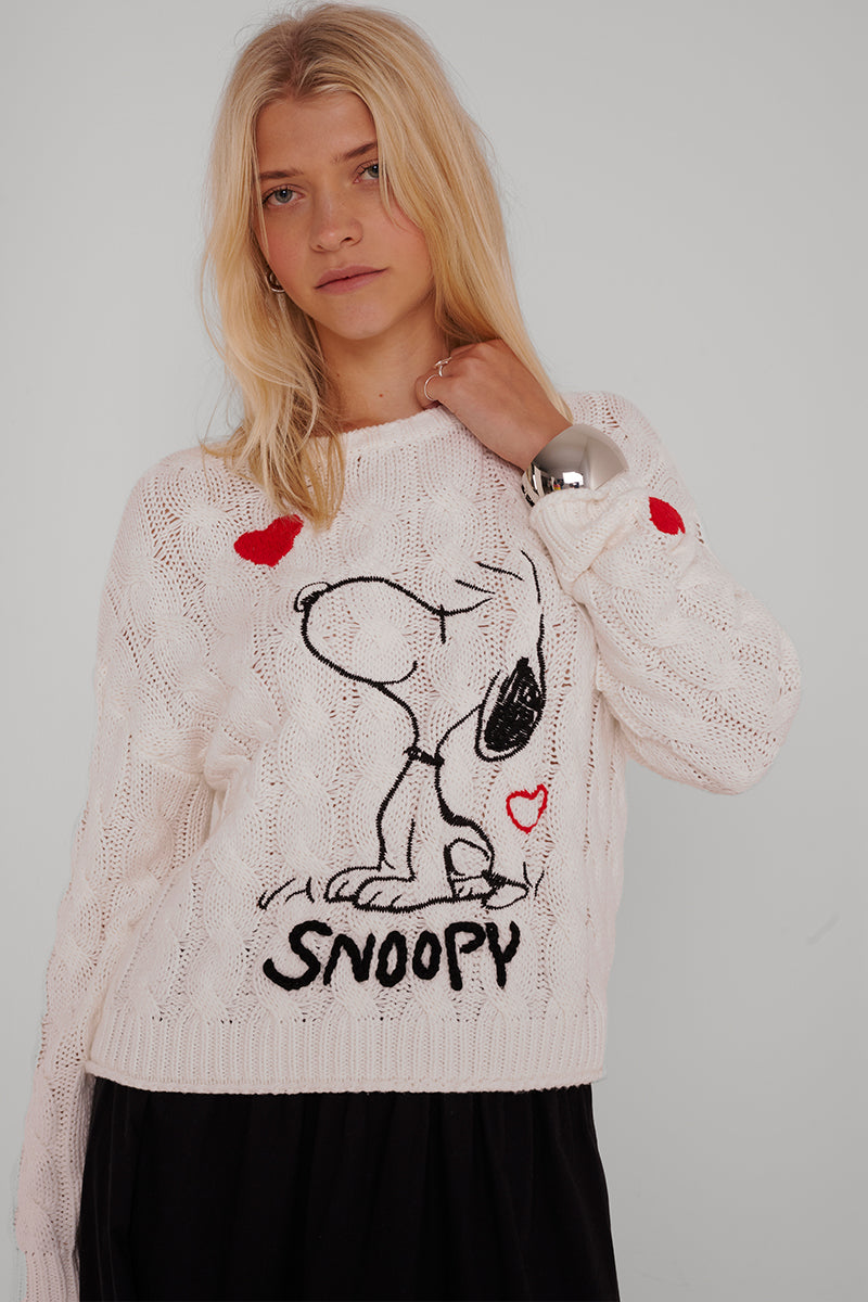Eine Frau mit langen blonden Haaren trägt einen weißen Baumwollmix-Zopfstrickpullover von Princess Goes Hollywood mit großer Snoopy-Stickerei, roten Herzen und dem Schriftzug "SNOOPY" darunter vor grauem Hintergrund.