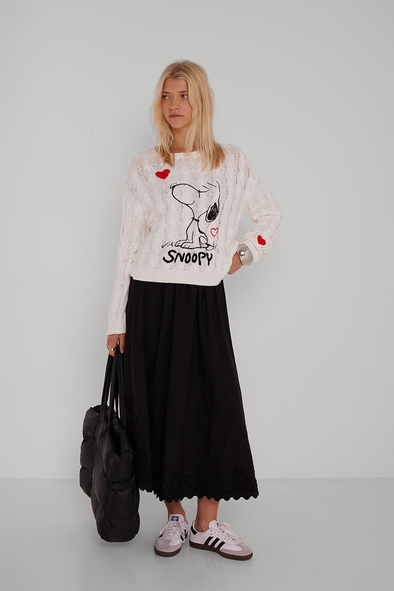 Eine junge Frau mit langen blonden Haaren trägt einen weißen Princess Goes Hollywood Snoopy-Pullover mit roter Herzstickerei, einen langen schwarzen Midirock und weiße Adidas-Turnschuhe. Sie trägt eine große schwarze Tasche und steht vor einer schlichten hellgrauen Wand und schaut nach links.