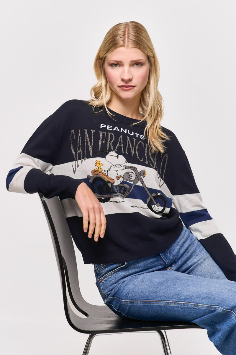 Eine Frau mit langen blonden Haaren sitzt auf einem schwarzen Stuhl und trägt einen marineblauen Princess Goes Hollywood Peanuts San Francisco Baumwoll-Kaschmir-Pullover mit einem Snoopy-auf-Motorrad-Motiv, perfekt kombiniert mit hellblauen Jeans.