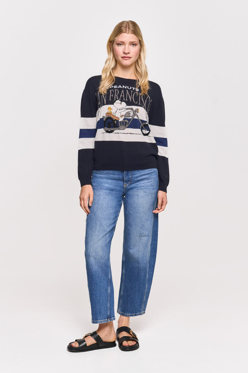 Eine blonde Frau in einem marineblauen Princess Goes Hollywood Peanuts San Francisco-Sweatshirt mit Snoopy-Motiv, blauen Jeans mit geradem Bein und schwarzen Sandalen vor einem schlichten weißen Hintergrund blickt nach vorne.