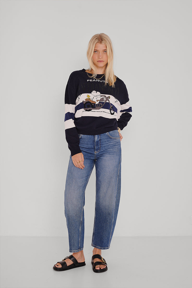 Eine junge Frau mit blondem Haar steht vor einer weißen Wand. Sie trägt ein marineblaues Princess Goes Hollywood Peanuts San Francisco Sweatshirt mit Snoopy, lässige Blue Jeans und schwarze Sandalen. Eine Hand steckt in ihrer Tasche und sie hat einen neutralen Gesichtsausdruck.