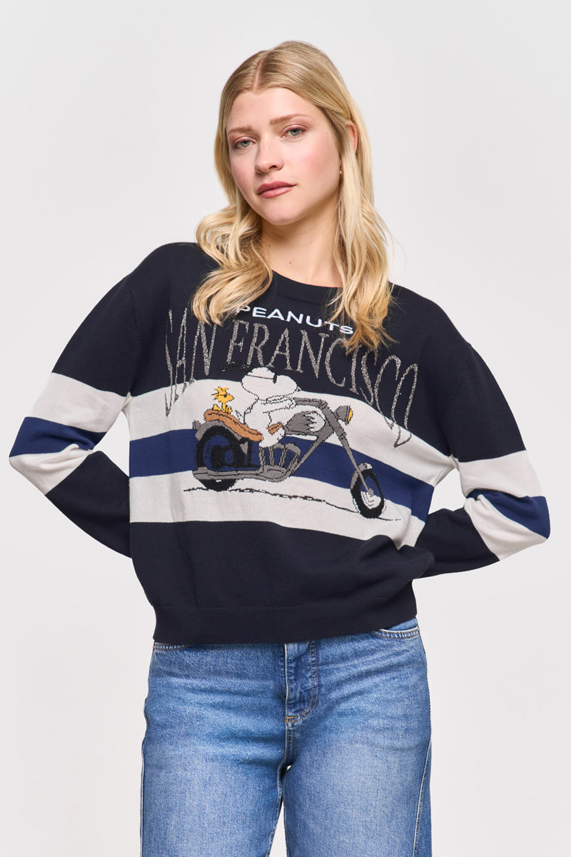 Eine blonde Frau in einem Princess Goes Hollywood-Pullover aus Baumwolle und Kaschmir mit einem Snoopy-Motiv und dem Schriftzug "Peanuts San Francisco" steht mit ausgebreiteten Armen, blauen Jeans und einem neutralen Gesichtsausdruck vor einem schlichten weißen Hintergrund.