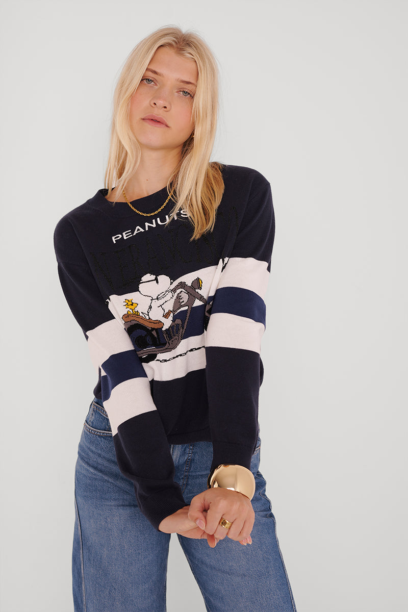 Eine blonde Frau trägt einen marineblauen und weißen Baumwoll-Kaschmir-Pullover mit Snoopy-Motiv und "PEANUTS"-Schriftzug, dazu Bluejeans und ein klobiges goldenes Armband, vor einem unifarbenen Hintergrund.