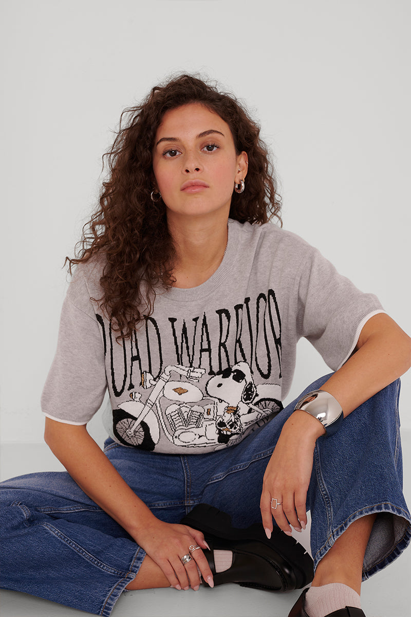 Eine junge Frau mit lockigem braunem Haar sitzt im Schneidersitz in blauen Jeans und einem hellgrauen Princess Goes Hollywood Road Warrior Pullover mit einem Snoopy-auf-Motorrad-Motiv. Sie trägt klobige Ohrringe und ein Armband und behält einen neutralen Gesichtsausdruck bei.