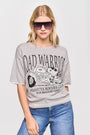 Eine Frau mit blondem Haar trägt eine große quadratische Sonnenbrille, blaue Jeans und ein lockeres Princess Goes Hollywood Snoopy-Motiv-T-Shirt mit der Aufschrift "ROAD WARRIOR PEANUTS BIKERS CLUB FOR BEAGLES ONLY" vor einem schlichten weißen Hintergrund.